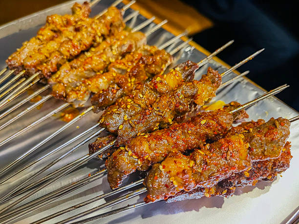 A close-up of several rows of Yang Rou Chuan (羊肉串), a popular and iconic street food originating from the Xinjiang region of China. These mutton or lamb skewers are expertly grilled over charcoal, giving them a distinct charred and smoky aroma. Each skewer consists of small, succulent pieces of meat seasoned with a vibrant blend of traditional spices, most notably toasted cumin seeds, coarse chili flakes, and salt.
The meat often includes small bits of fat that render during the grilling process, making the skewers incredibly juicy and flavorful. The skewers are typically prepared on long, thin metal needles, as seen in the image, which allow them to be rotated easily over high-heat grills. This dish is a staple of Northeastern Chinese BBQ and is frequently enjoyed as a savory snack or a hearty meal accompanied by cold beer.
